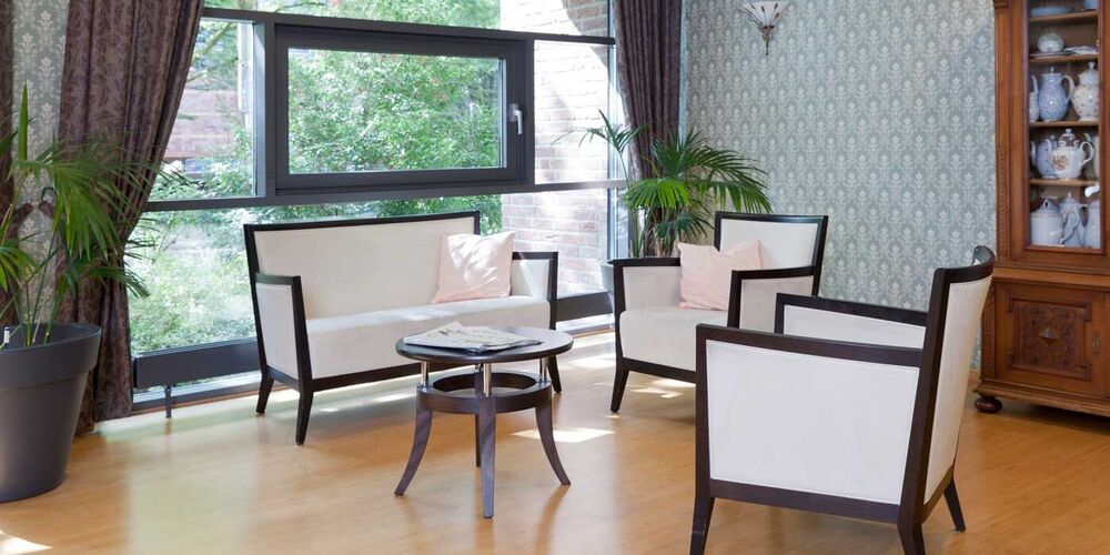 Einrichtung eines Salons im Haus Maria Hilf. In diesem steht ein Sofa und zwei Sessel mit einem kleinen Tisch. Außerdem befindet sich ein Schrank mit Geschirr in diesem.