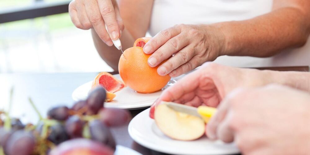 Zwei Hände die einen Apfel schneiden und zwei weitere schneiden eine Grapefruit. 