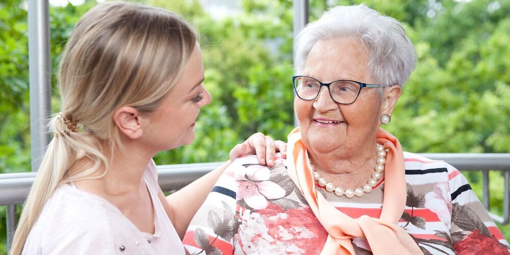 Eine Seniorin und eine junge Frau schauen sich an. Sie halten eine Hand und die junge Frau hat ihre andere Hand auf der Schulter der Seniorin.